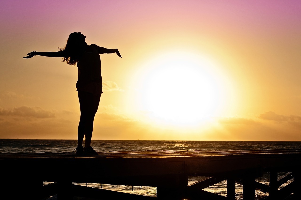 Mujer con los brazos abiertos frente al mar al atardecer, simbolizando bienestar, libertad y equilibrio emocional.